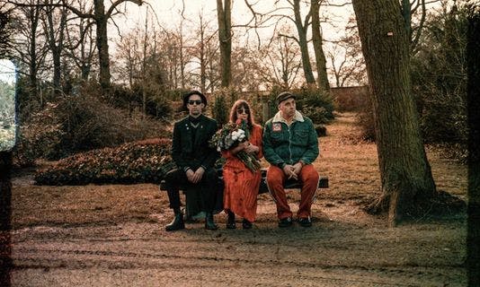 Die drei Bandmitglieder sitzen auf einer Bank in einem herbstlichen Park und tragen Sonnenbrillen. Die Sängerin hält einen Blumenstrauß in der Hand.